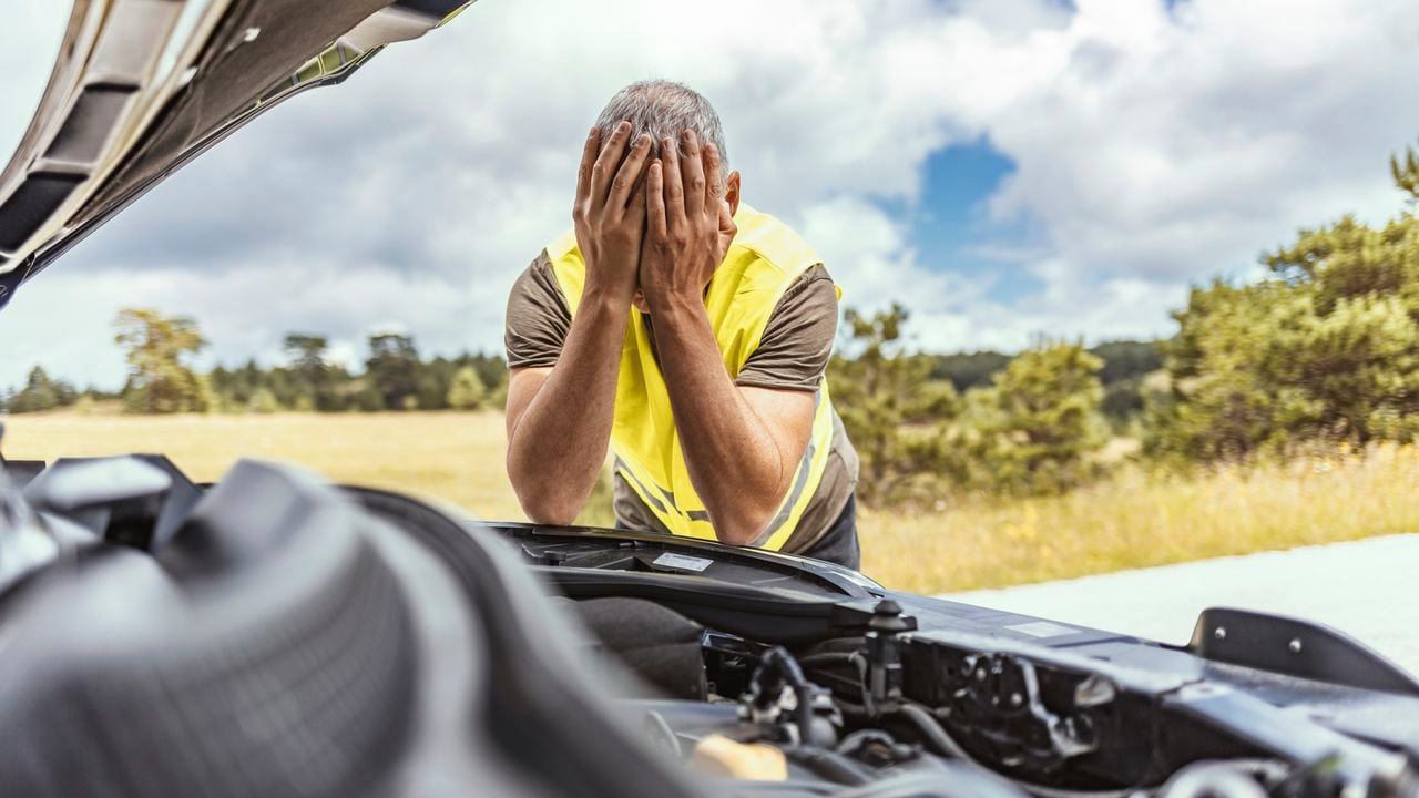 Worried mature man holding his head by hands standing near his broken old car with raised hood on the roadside of a rural road