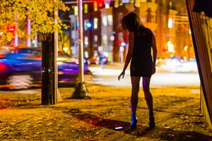 A young women alone on a street corner in a big city at night, with a dropped cell phone on the ground.