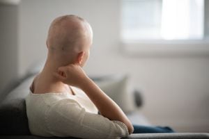 Una mujer está dentro de su sala de estar. Su cabeza está afeitada debido a la quimioterapia. Ella está sentada y luciendo pensativa.