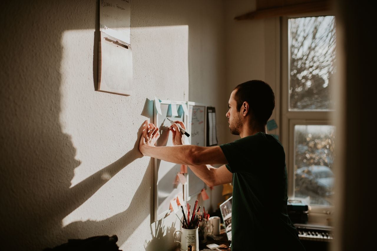 A young man, who is clearly organised in nature, writes reminders and affirmations on a white board on his wall, within a small home office environment. Sunshine pours through the window creating shadows on his wall, which provides a space for copy.