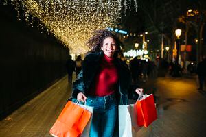 Cheerful young Latin American woman, carrying shopping bags and running on a christmas illuminated street, at night.