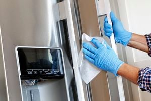A close up of a Hispanic man wearing disposable gloves as he cleans a stainless steel refrigerator door handle with a disinfectant wipe.