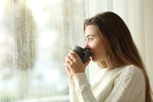 Retrato lateral de un adolescente relajado tomando café mirando afuera a través de una ventana en un día lluvioso de invierno en casa