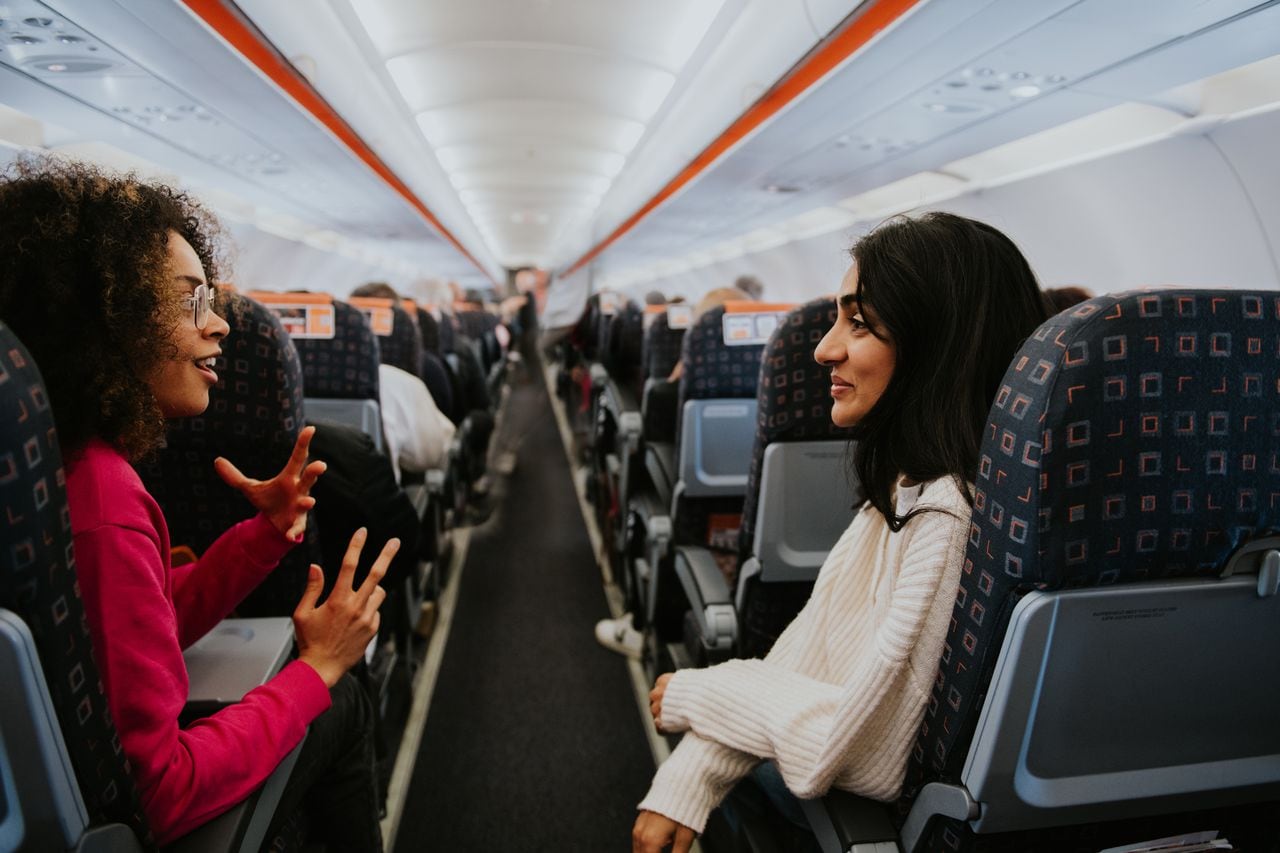 Two young woman sit on a commercial aircraft. They turn their bodies towards each other and over the aisle to have a chat. They are happy and engaged in a friendly conversation.