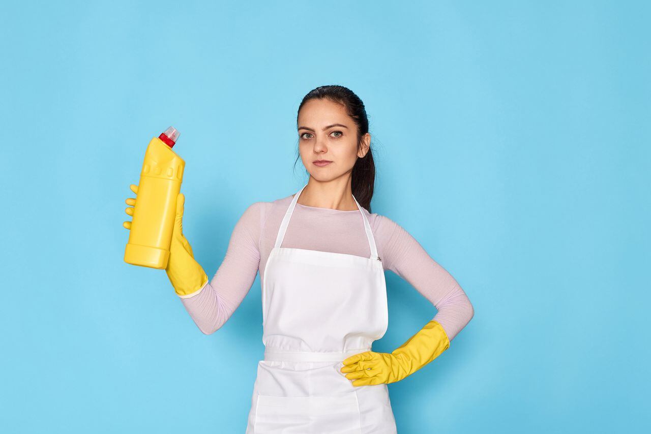 beautiful caucasian woman in yellow rubber gloves and cleaner apron holding bottle of detergent on blue background.