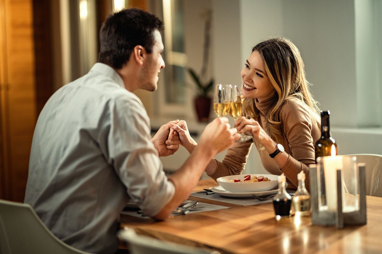 Mujer feliz y su novio tomados de la mano mientras brindaban con champán mientras cenaban en la mesa del comedor.
