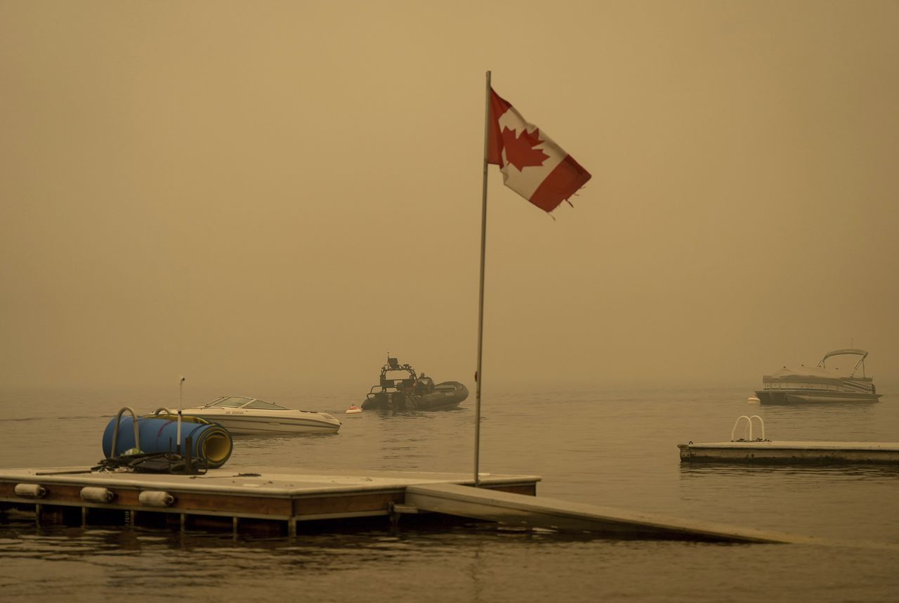 El humo espeso del incendio forestal del lago Lower East Adams llena el aire alrededor de una bandera canadiense ondeando en el viento mientras los oficiales de la Real Policía Montada de Canadá en un barco patrullan el lago Shuswap, en Scotch Creek, Columbia Británica, el domingo 20 de agosto de 2023