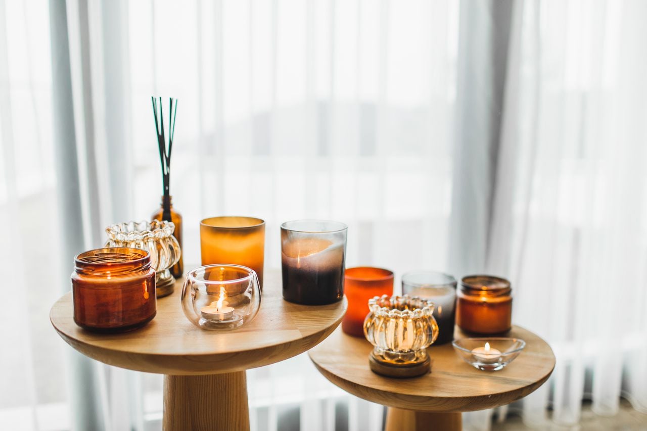 Organic soya wax candles in different glass candlestick in brown bowls on wooden tea-table near comfortable armchair. Cosy, comfortable and soft home background. Hygge and aromatherapy concept.