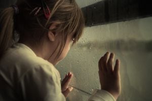 Sad little girl looking through the rainy window. Grain added for texture.