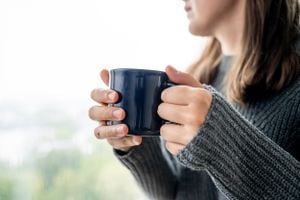 Retrato de una joven sonriente en suéter con taza de café. Ella toma café sentada cerca de la ventana, llueve afuera