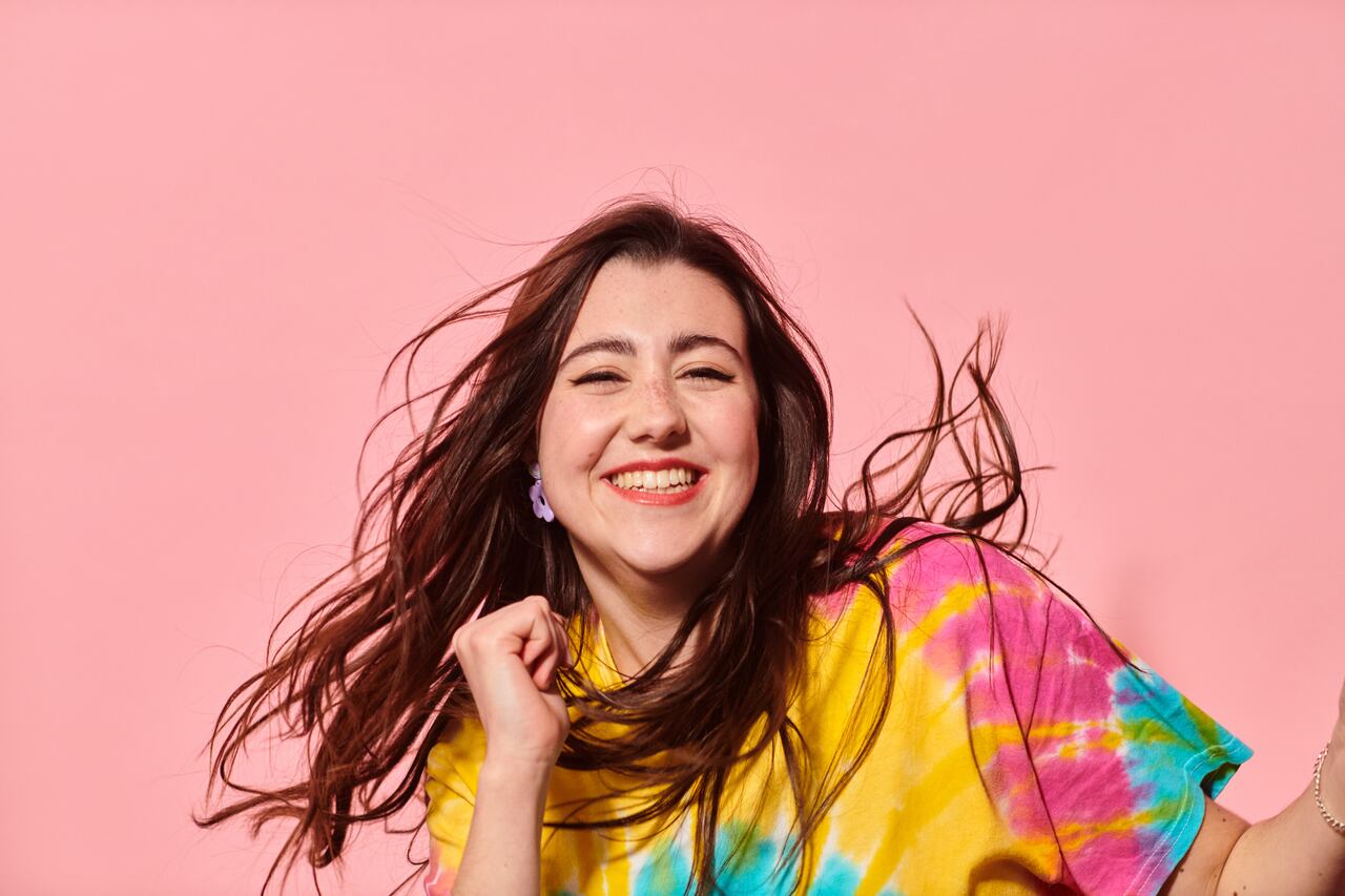 Close up of happy young woman with profound deafness in colourful tie-dye top against pink background laughing and dancing