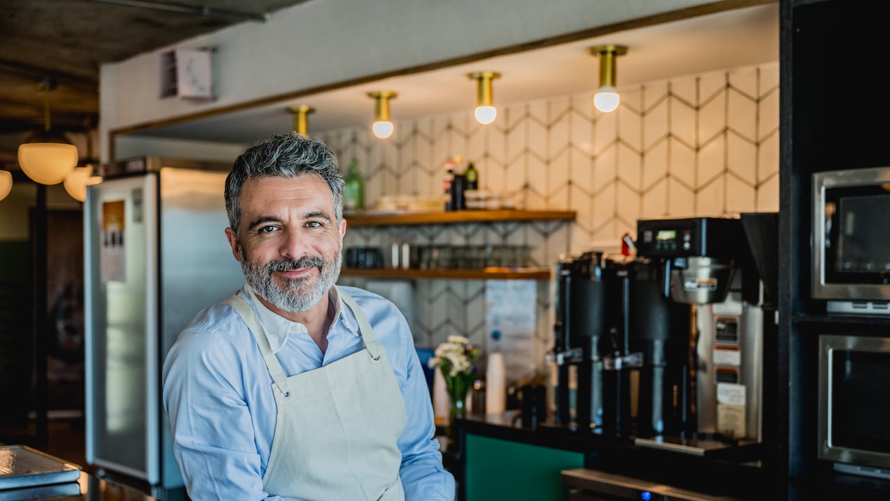 Barista barbudo vistiendo delantal y apoyado en el mostrador de la cafetería corporativa en la oficina de Buenos Aires y sonriendo a la cámara.