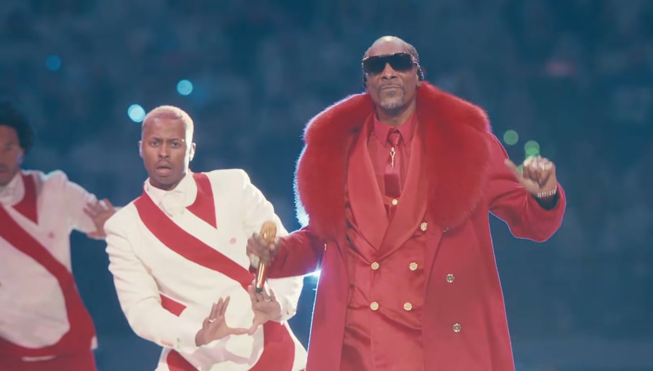 Snoop Dogg encabeza el “Holiday Halftime Party” de la NFL, vistiendo un traje rojo navideño frente a una orquesta y bailarines en el U.S. Bank Stadium de Minneapolis.