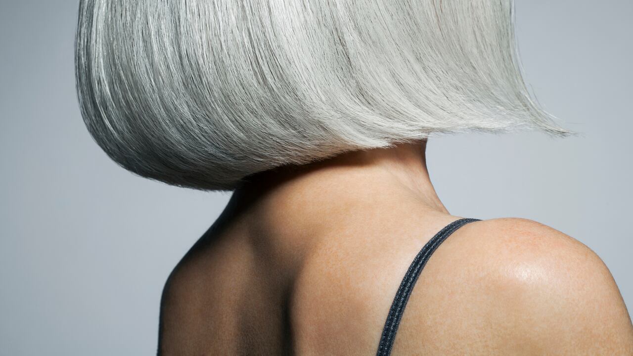 Mature woman with silvery, grey hair styled in a chin length bob in front of a grey background wearing a spaghetti strap tank top turning away from camera, cropped.