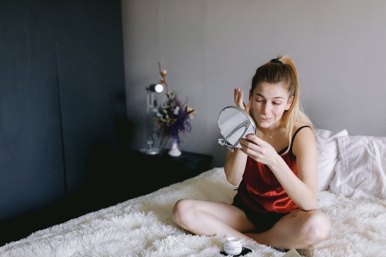 High angle of young female in sleepwear looking at mirror and spreading cream on face while sitting cross legged on bed during skin care routine at home