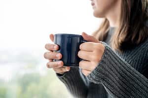 Retrato de una joven sonriente en suéter con taza de café. Ella toma café sentada cerca de la ventana, llueve afuera