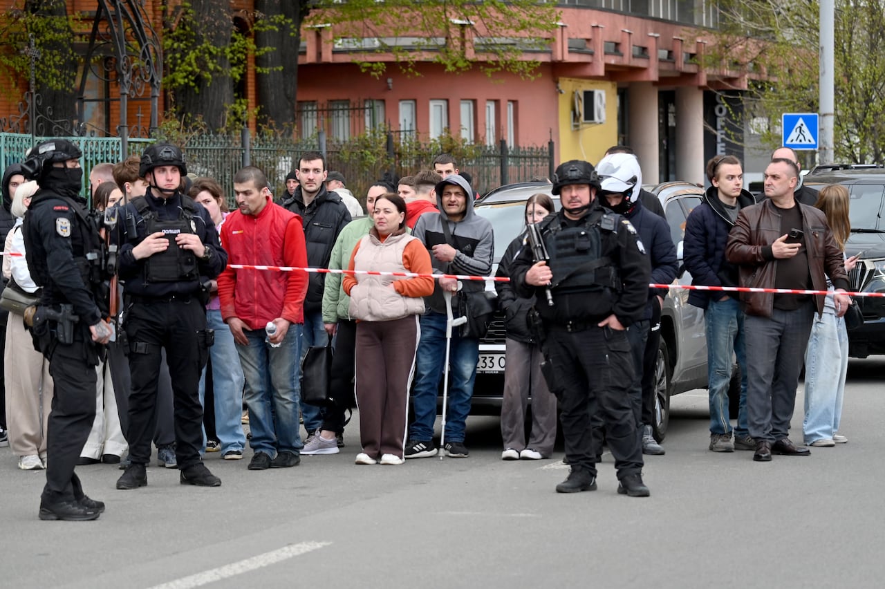 People stand behind police officers outside a supermarket following a shooting in Kyiv on April 18, 2026. A gunman opened fire in Kyiv on April 18, 2026, killing at least five people, wounding others and taking hostages at a supermarket before being killed during an arrest attempt, officials said. Ukrainian President said that at least five people were dead and 10 hospitalised with wounds and trauma after the shooting in a residential district of the capital. (Photo by Sergei SUPINSKY / AFP)