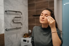 woman wiping face with moisturizing lotion with cotton pad in front of mirror in bathroom at home