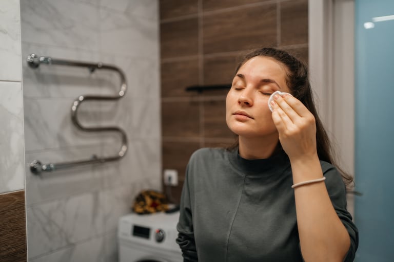 woman wiping face with moisturizing lotion with cotton pad in front of mirror in bathroom at home