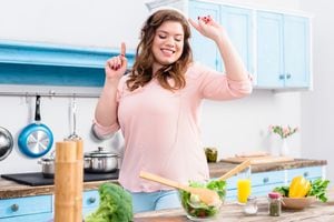 Alegre mujer con sobrepeso escuchando música en auriculares y bailando en la mesa con verduras frescas en la cocina de casa